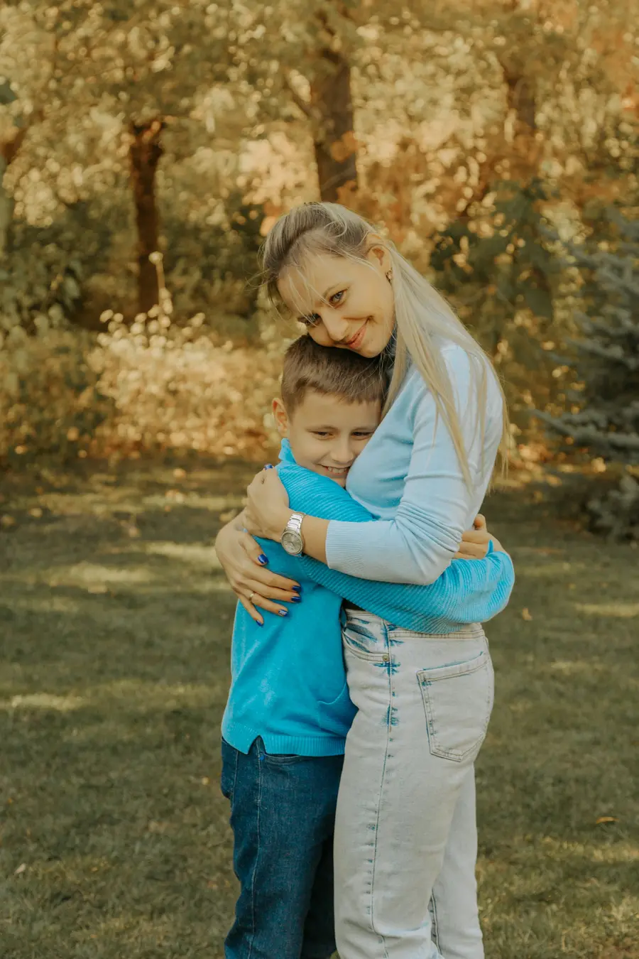 Mãe sorridente abraçando o filho pequeno ao ar livre num parque claro, céu azul e