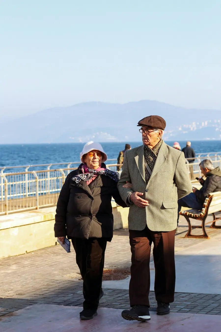 Casal de uns 65 anos sorrindo em calçadão à beira-mar em dia de céu azul