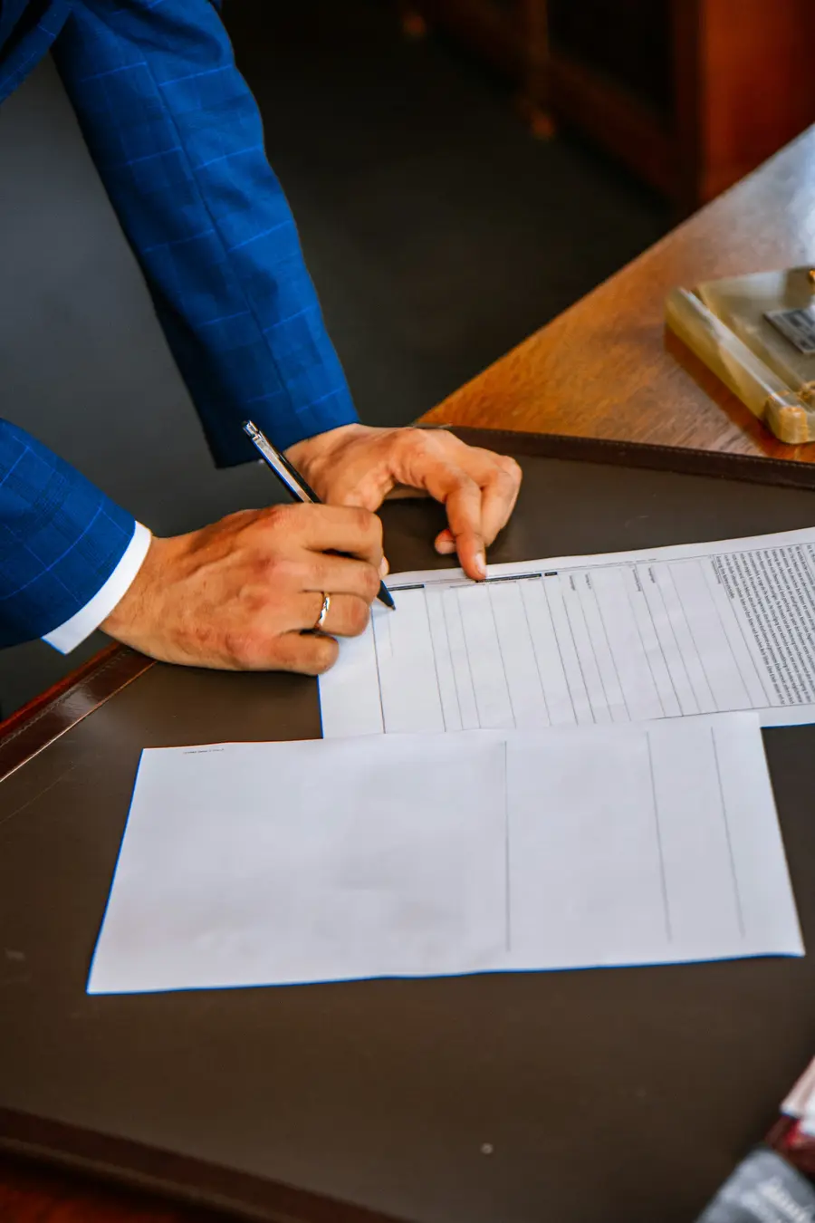 Empresário sorridente assinando um documento numa mesa clara, com notebook ao lado e detalhes em azul