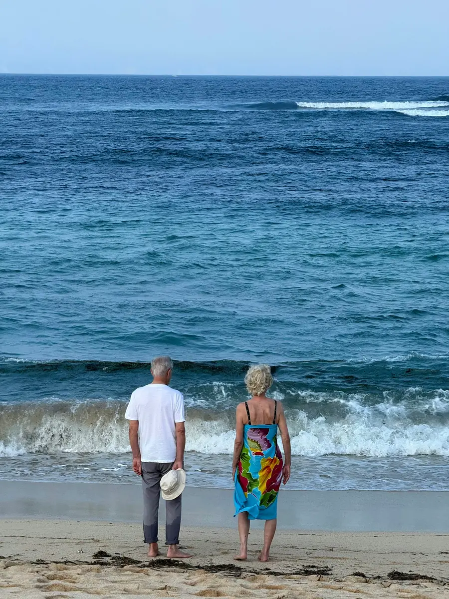 Casal de uns 60 anos sorrindo em caminhada na orla, céu azul e mar ao fundo