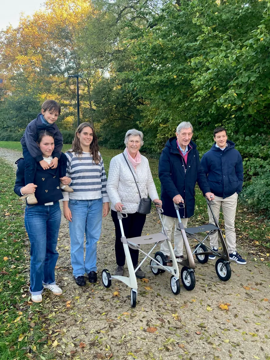 Família sorridente caminhando em parque arborizado num dia claro, tons de verde e azul