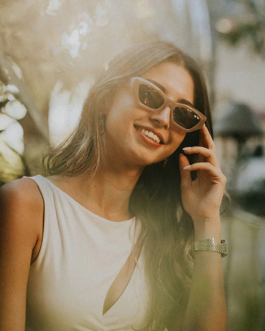 Mulher de uns 30 anos sorrindo enquanto olha o celular numa rua arborizada e ensolarada