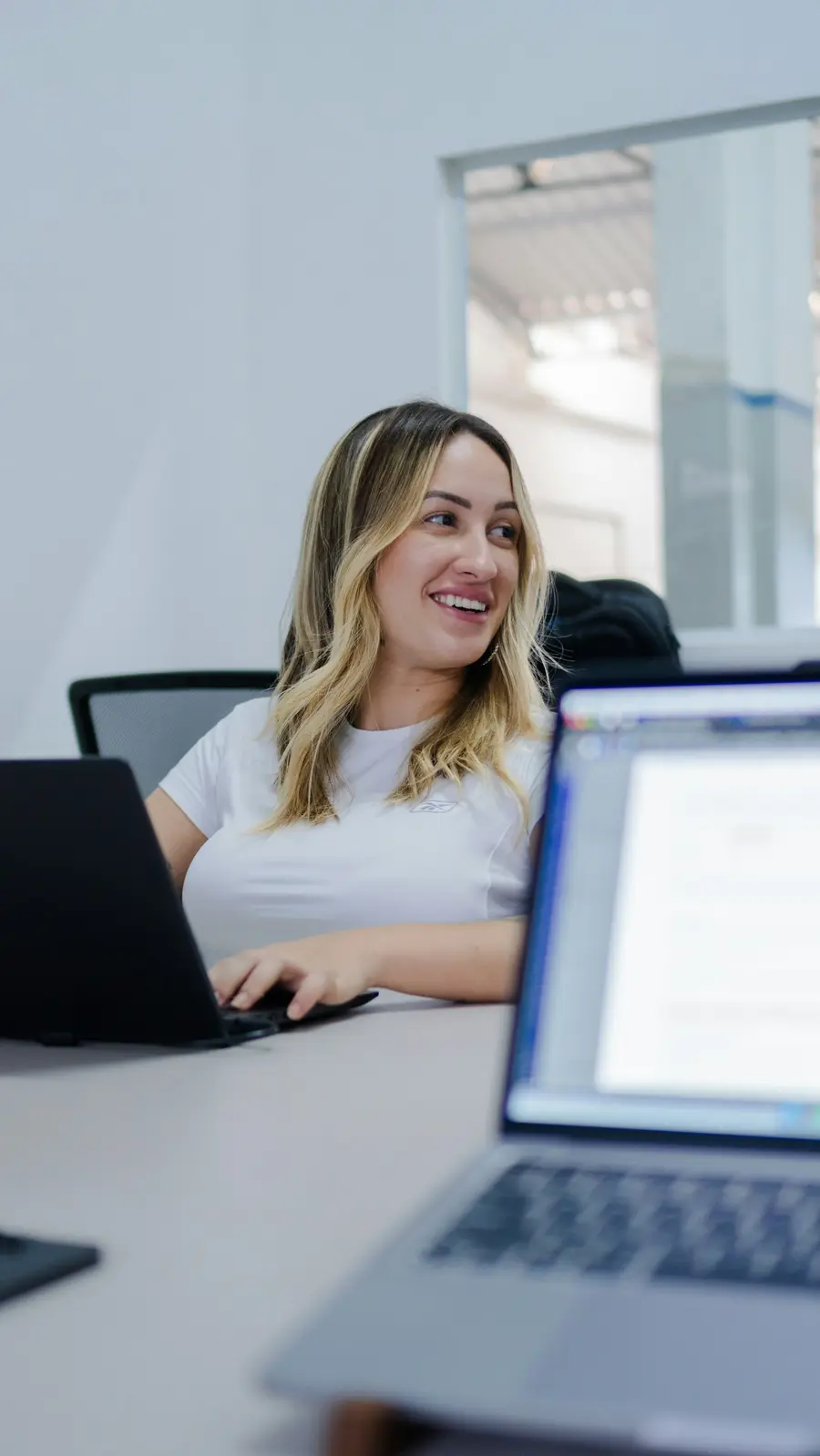 Mulher de uns 35 anos sorrindo enquanto lê no notebook em escritório claro