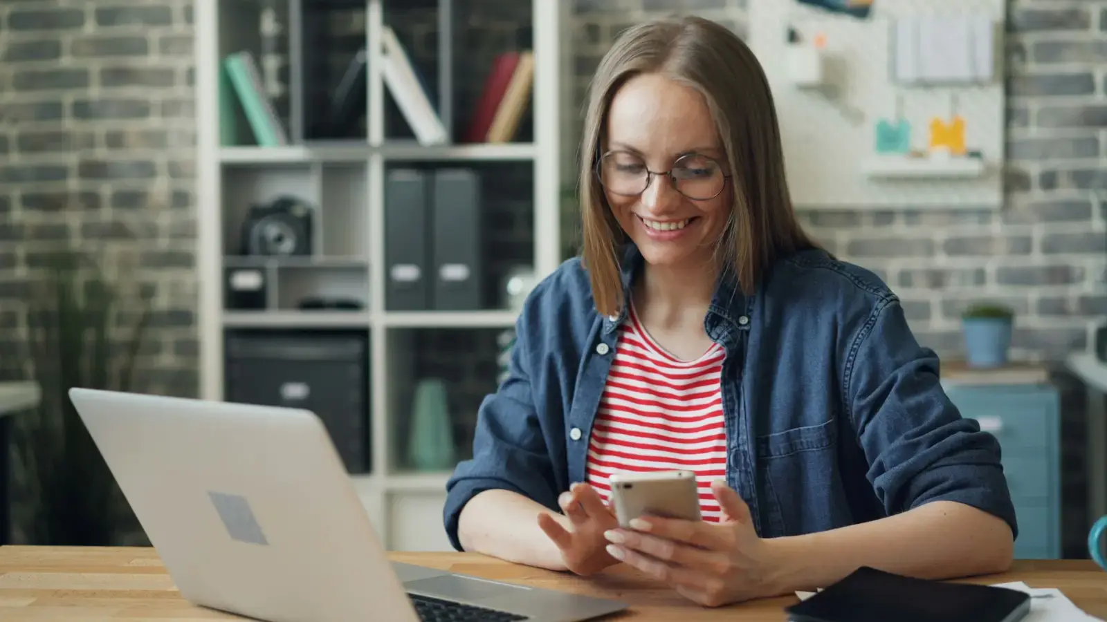 Mulher sorrindo ao consultar o celular diante do notebook